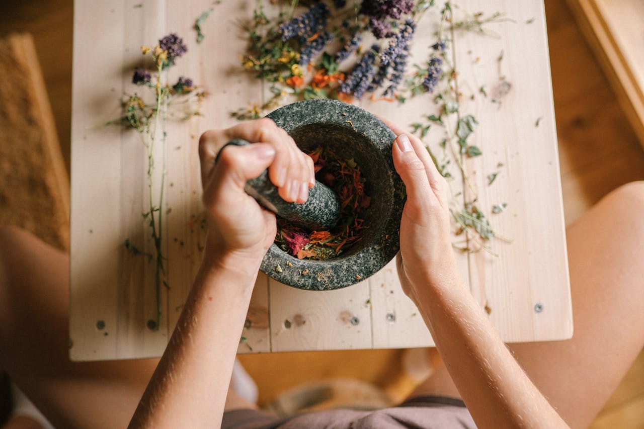 Top view of hands grinding dried herbs and petals with a mortar and pestle on a wooden table.